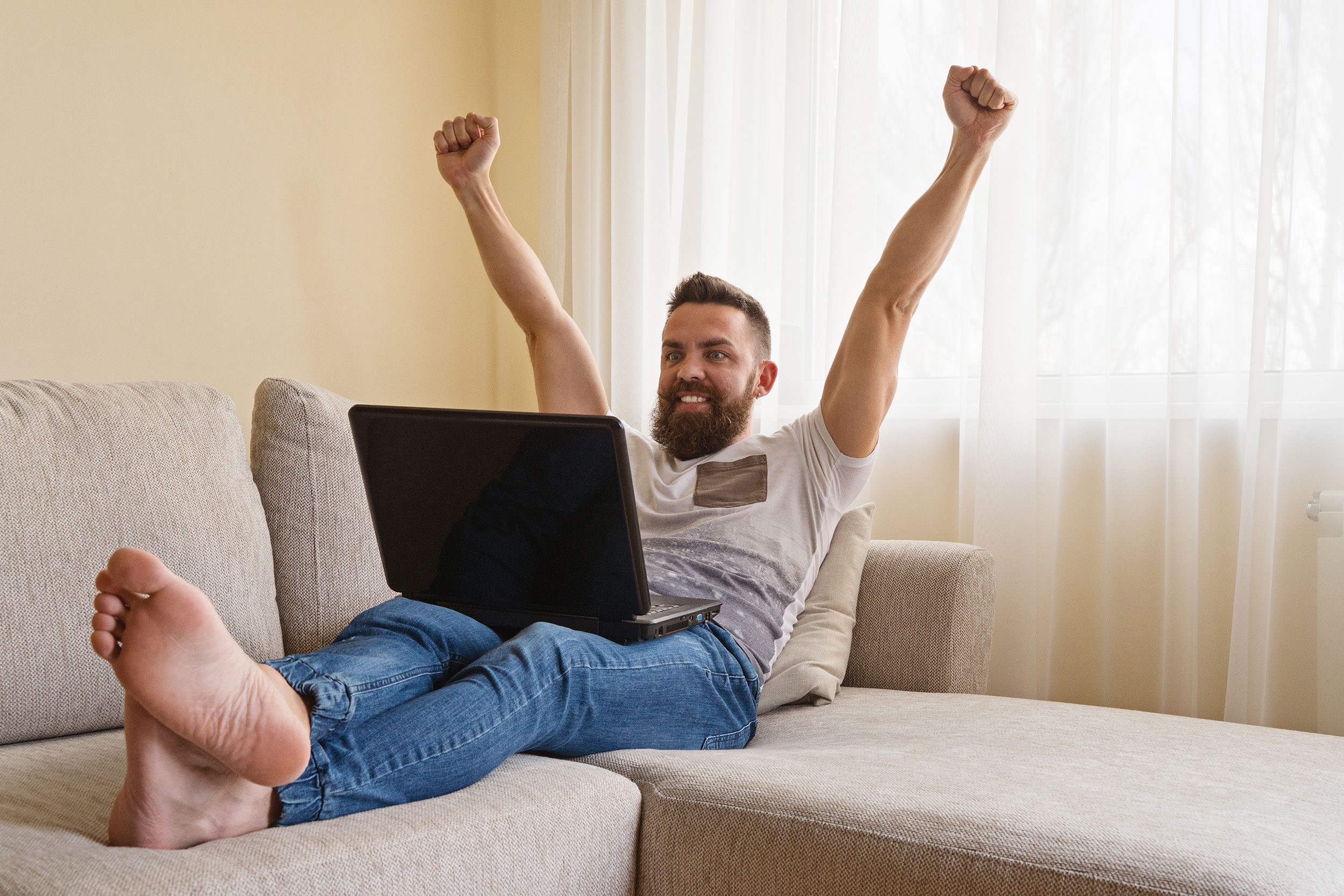 Man with computer raising hands in victory on a couch representing a freelance web designer who is hitting 6 figures