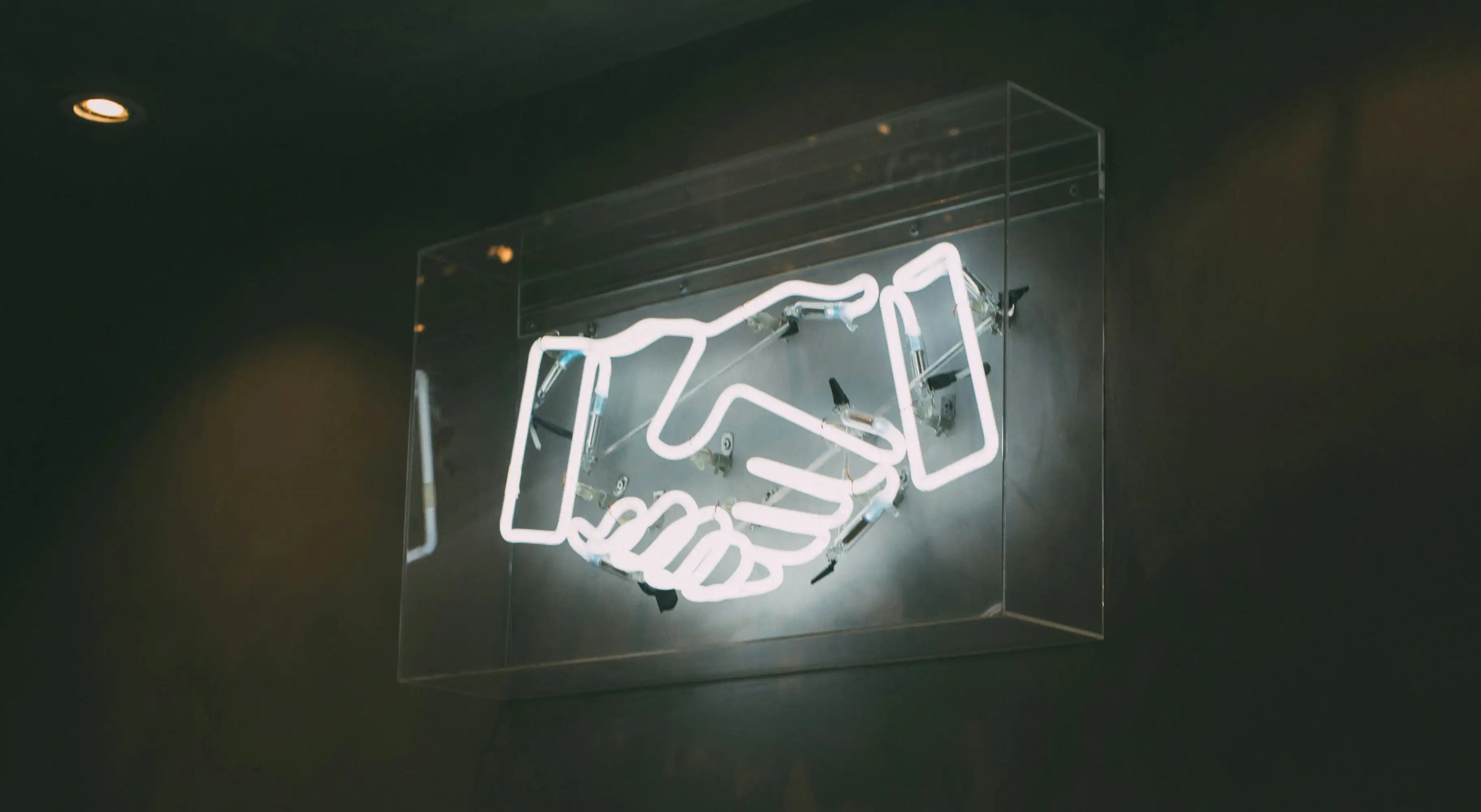 A neon sign of two hands shaking, symbolizing a business deal or partnership, mounted on a dark wall. The image represents the concept of networking and making connections—key strategies in ‘How to Get Your First Web Design Client.